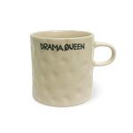 A beige ceramic mug with a textured surface and a handle, showing the text "DRAMA QUEEN" in black capital letters, with the "Q" stylized to include a small crown, set against a white background.