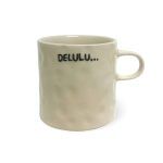 A beige ceramic mug with a slightly textured surface and a handle, featuring the text "DELULU..." written in black capital letters with three dots, set against a white background.