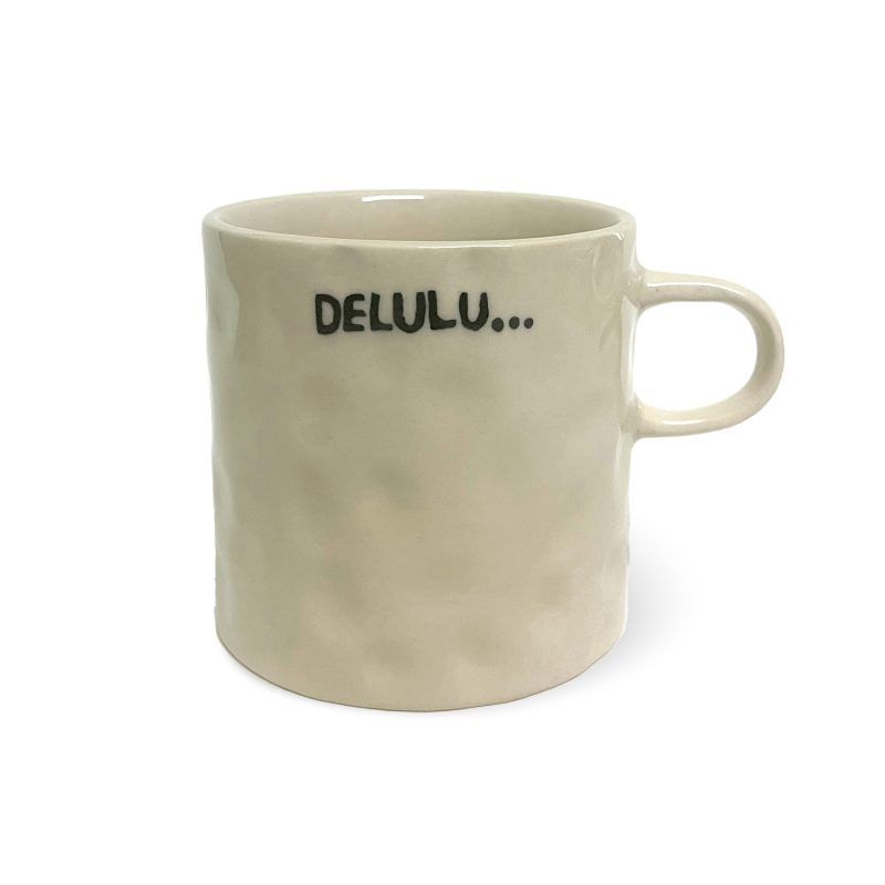 A beige ceramic mug with a slightly textured surface and a handle, featuring the text "DELULU..." written in black capital letters with three dots, set against a white background.