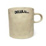 white background image of an off-white, textured ceramic mug. The mug has the word “DELULU…” printed in bold black letters.