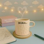Off-white ceramic coffee mug with 'DELULU...' text, on a pastel-themed desk with books, heart-shaped steam, and dreamy decor.
