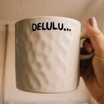 An off-white, textured ceramic mug held by a hand with light purple nail polish. The mug has the word “DELULU…” printed in bold black letters.