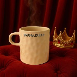 Off-white ceramic coffee mug with 'DRAMA QUEEN' text, placed on red velvet with a golden crown in the background.