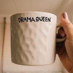 An off-white, textured ceramic mug held by a hand with light purple nail polish. The mug features the phrase “DRAMA QUEEN” in bold black letters, with a stylized “Q.”