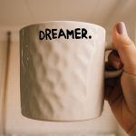 An off-white, textured ceramic mug held by a hand with light purple nail polish. The mug is labeled with the word “DREAMER.” in bold black text, ending with a period.