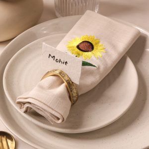 A close-up of a beige cloth napkin with a sunflower embroidery, neatly folded and held by a textured antique gold napkin ring. A name card reading "Mohit" is tucked into the ring, placed on a stacked set of speckled ceramic plates.