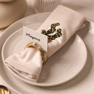 A beige napkin featuring a cactus embroidery is neatly rolled and held by an antique gold napkin ring. A name card labeled "Mayank" is tucked into the ring, all placed on a set of speckled ceramic plates.