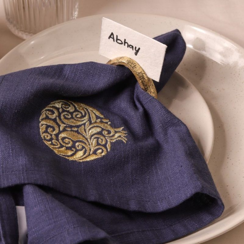 A dark blue napkin with an intricate gold embroidered motif is partially tucked into an antique gold napkin ring. A name card labeled "Abhay" is inserted into the ring, and the setting rests on speckled ceramic plates.