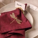 A maroon napkin with a detailed gold embroidered dragonfly is partially tucked into an antique gold napkin ring. A name card labeled "Ayushi" is inserted in the ring, all placed on speckled ceramic plates.