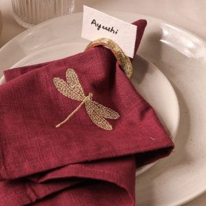 A maroon napkin with a detailed gold embroidered dragonfly is partially tucked into an antique gold napkin ring. A name card labeled "Ayushi" is inserted in the ring, all placed on speckled ceramic plates.