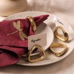 A dragonfly-embroidered maroon napkin is secured with an antique gold napkin ring and rests on stacked ceramic plates. Three additional antique gold napkin rings are arranged nearby, with name cards labeled "Ayushi" and "Sneha."