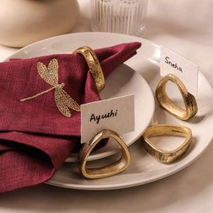 A dragonfly-embroidered maroon napkin is secured with an antique gold napkin ring and rests on stacked ceramic plates. Three additional antique gold napkin rings are arranged nearby, with name cards labeled "Ayushi" and "Sneha."