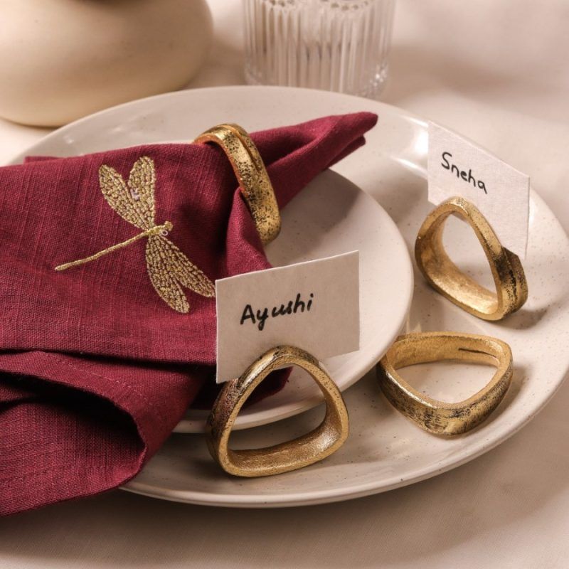 A dragonfly-embroidered maroon napkin is secured with an antique gold napkin ring and rests on stacked ceramic plates. Three additional antique gold napkin rings are arranged nearby, with name cards labeled "Ayushi" and "Sneha."