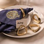 A gold-pomegranate embroidered navy blue napkin is secured with an antique gold napkin ring and placed on stacked ceramic plates. Three more antique gold napkin rings are displayed nearby, two of which hold name cards labeled "Abhay" and "Sneha."