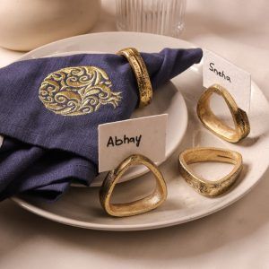 A gold-pomegranate embroidered navy blue napkin is secured with an antique gold napkin ring and placed on stacked ceramic plates. Three more antique gold napkin rings are displayed nearby, two of which hold name cards labeled "Abhay" and "Sneha."