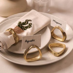 A cactus-embroidered beige napkin is secured with an antique gold napkin ring on stacked ceramic plates. Three additional antique gold napkin rings are arranged on the plate, two holding name cards labeled "Ayushi" and "Mayank."