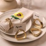 A beige napkin with sunflower embroidery and antique gold napkin ring rests on a ceramic plate. Three additional gold napkin rings are placed nearby, holding name cards labeled "Ayushi," "Ankit," and one empty ring.