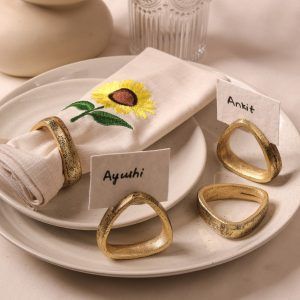 A beige napkin with sunflower embroidery and antique gold napkin ring rests on a ceramic plate. Three additional gold napkin rings are placed nearby, holding name cards labeled "Ayushi," "Ankit," and one empty ring.
