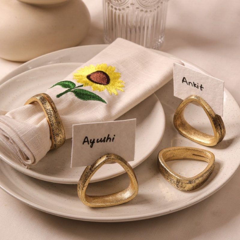 A beige napkin with sunflower embroidery and antique gold napkin ring rests on a ceramic plate. Three additional gold napkin rings are placed nearby, holding name cards labeled "Ayushi," "Ankit," and one empty ring.