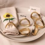 A sunflower-embroidered beige napkin is held by an antique gold napkin ring on a ceramic plate. Surrounding it are five triangular antique gold napkin rings, two of which hold name cards labeled "Abhay" and "Ankit."