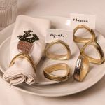 A cactus-embroidered napkin with an antique gold napkin ring is placed on ceramic plates. Five other antique gold napkin rings surround the napkin, two displaying name cards reading "Ayushi" and "Mayank."