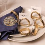 A dark blue napkin with gold pomegranate embroidery is styled with an antique gold napkin ring and set on ceramic plates. Five additional antique gold napkin rings are arranged around it, two holding name cards labeled "Mohit" and "Mayank."