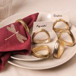 A maroon napkin with gold dragonfly embroidery is styled with an antique gold napkin ring and placed on ceramic plates. Five more antique gold napkin rings surround the setting, two holding name cards reading "Ayushi" and "Sneha."