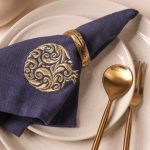 A navy blue napkin featuring a gold embroidered pomegranate design is held by an antique gold napkin ring and placed on speckled ceramic plates. Gold-toned cutlery - a spoon and a fork - is arranged beside the plate.