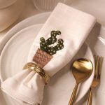 A beige napkin embroidered with a potted cactus design is elegantly rolled and held by an antique gold napkin ring. It rests on a set of speckled ceramic plates, accompanied by gold-toned cutlery including a spoon and a fork, arranged neatly on the right.