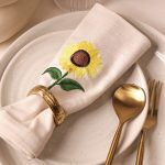 A sunflower-embroidered beige napkin is placed on a ceramic plate with an antique gold napkin ring. Next to the plate is a set of gold cutlery including a spoon and a fork, arranged neatly.