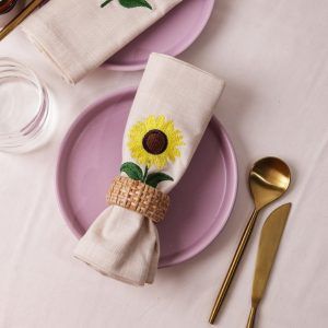 Two beige napkins with sunflower embroidery rest on mauve plates. One is held by a woven rattan napkin ring, while the other lies flat. Gold cutlery and a glass are arranged nearby on a light-colored tablecloth.