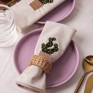 A beige napkin embroidered with a green cactus in a tan pot is rolled and secured with a woven rattan napkin ring, placed on a lilac ceramic plate. Gold-toned cutlery and a glass of water accompany the setup.