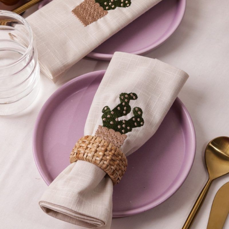 A beige napkin embroidered with a green cactus in a tan pot is rolled and secured with a woven rattan napkin ring, placed on a lilac ceramic plate. Gold-toned cutlery and a glass of water accompany the setup.