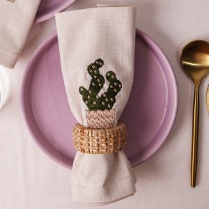 A close-up of a cactus-embroidered beige napkin held by a woven rattan napkin ring, centered on a lilac plate. The setting is complemented by a gold spoon and knife on the side.