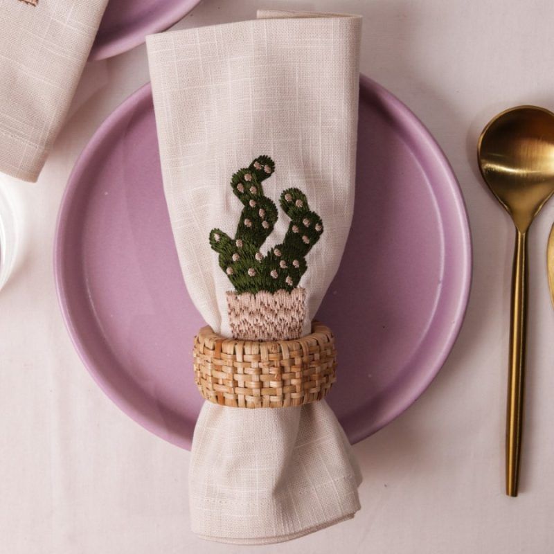 A close-up of a cactus-embroidered beige napkin held by a woven rattan napkin ring, centered on a lilac plate. The setting is complemented by a gold spoon and knife on the side.