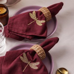 Two maroon napkins with detailed gold dragonfly embroidery are each secured with woven rattan napkin rings and placed on lilac ceramic plates. A glass and a candle are partially visible on the table.