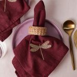 A single maroon napkin with gold dragonfly embroidery is elegantly rolled and held with a woven rattan napkin ring, resting on a lilac ceramic plate. Gold-toned cutlery is arranged on the side.