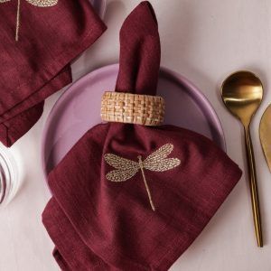 A single maroon napkin with gold dragonfly embroidery is elegantly rolled and held with a woven rattan napkin ring, resting on a lilac ceramic plate. Gold-toned cutlery is arranged on the side.