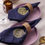 Two navy blue napkins with intricate gold embroidered pomegranate motifs are each rolled and secured with woven rattan napkin rings, placed on mauve ceramic plates. A lit amber candle, water glass, and gold-toned cutlery complete the elegant table setting.