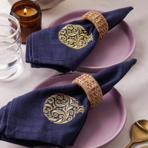 Two navy blue napkins with intricate gold embroidered pomegranate motifs are each rolled and secured with woven rattan napkin rings, placed on mauve ceramic plates. A lit amber candle, water glass, and gold-toned cutlery complete the elegant table setting.