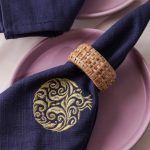 A close-up of a navy blue napkin with a gold embroidered pomegranate motif, held by a woven rattan napkin ring and placed on a mauve ceramic plate. The texture of the embroidery and weave are clearly visible.