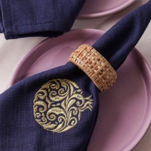 A close-up of a navy blue napkin with a gold embroidered pomegranate motif, held by a woven rattan napkin ring and placed on a mauve ceramic plate. The texture of the embroidery and weave are clearly visible.