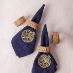 Two rolled navy blue napkins with gold pomegranate embroidery are held by woven rattan napkin rings and displayed on a light background. Two extra rattan rings are scattered around them.