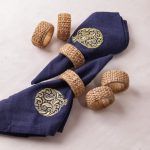 A pair of navy blue napkins with gold pomegranate embroidery are styled with woven rattan napkin rings and laid on a white surface. Four additional rattan rings are placed decoratively around them.