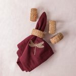 A maroon napkin embroidered with a gold dragonfly is styled with a woven rattan napkin ring and placed on a white surface. Three additional rattan napkin rings are scattered nearby.
