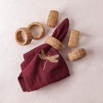 A maroon napkin featuring a gold dragonfly embroidery is held by a woven rattan napkin ring, displayed on a light background. Five woven napkin rings are arranged around it