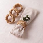 A rolled napkin with cactus embroidery is held by a woven rattan napkin ring and laid on a white surface. Three additional woven rattan napkin rings are arranged beside it.