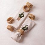 Two cactus-embroidered beige napkins - one rolled with a woven rattan napkin ring and the other folded - are displayed on a white background. Five woven napkin rings are scattered around them.
