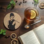 A cork coaster featuring Audrey Hepburn from Breakfast at Tiffany’s on a wooden table with a cup of tea and lemon, open book, dried citrus slices, mint leaves, and coffee beans.