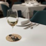 A cork coaster featuring Audrey Hepburn from Breakfast at Tiffany’s with a glass of white wine placed on top, set on a fine dining restaurant table with plate, napkin, and cutlery.